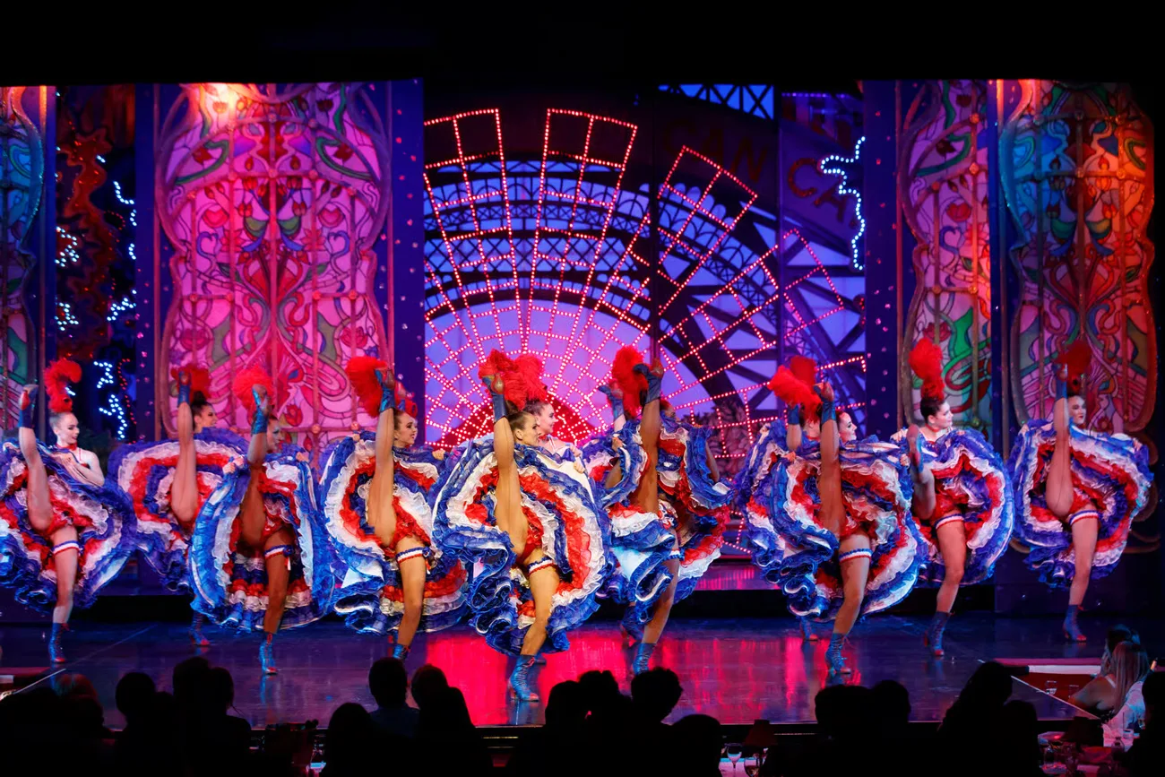 Féerie Can-can dancers in red, white, and blue ruffled skirts performing high kicks on a brightly lit stage in front of a seated audience.