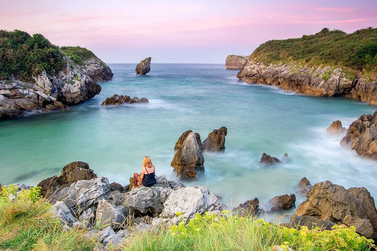 Woman sitting on coastal rocks overlooking a turquoise cove on Spain's northern coast at sunset